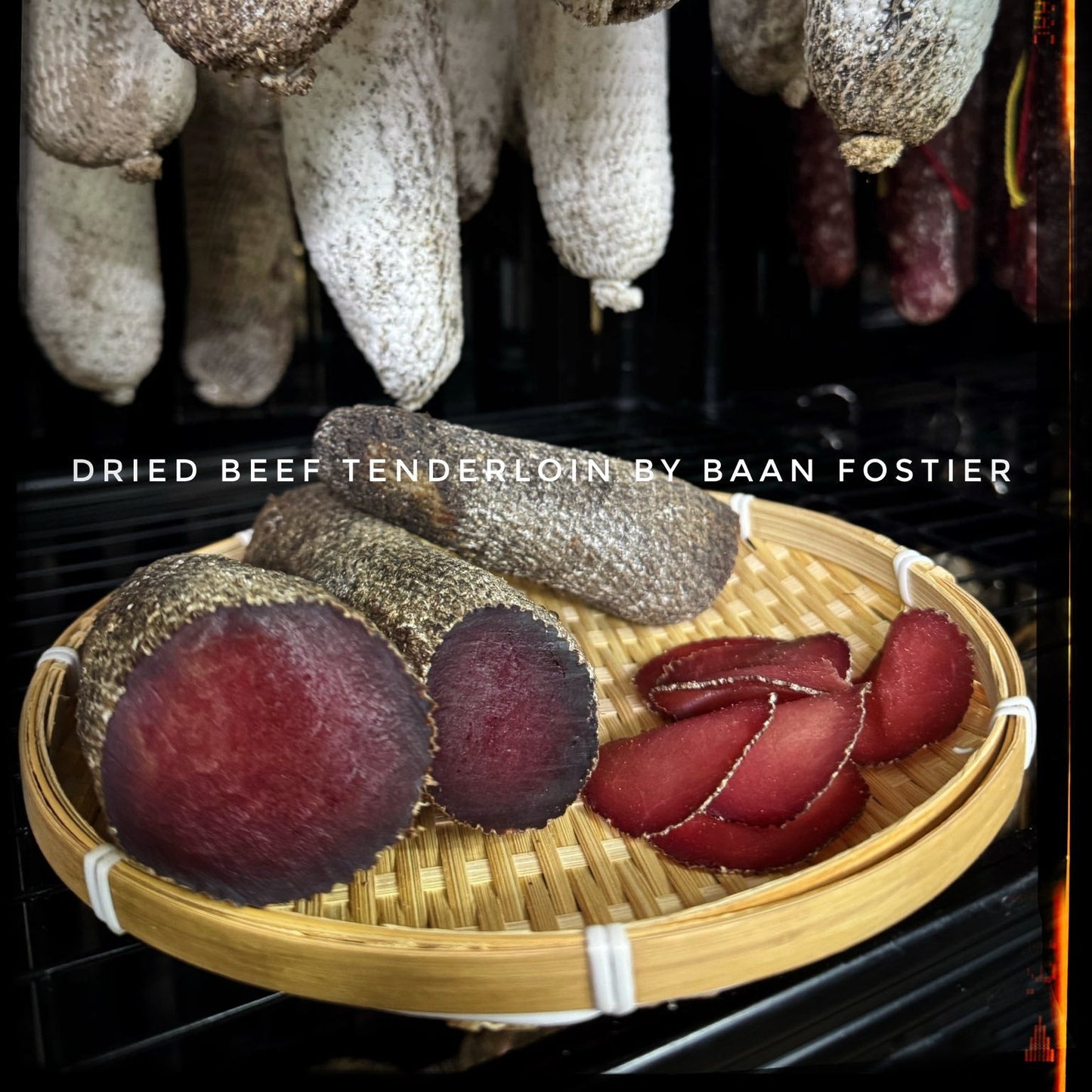 Dried beef tenderloin displayed on a woven tray with hanging dried beef in the background.